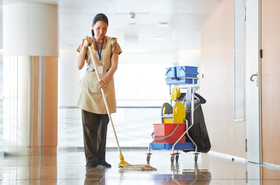 Adult cleaner maid woman with mop and uniform cleaning corridor pass or hall floor of business building